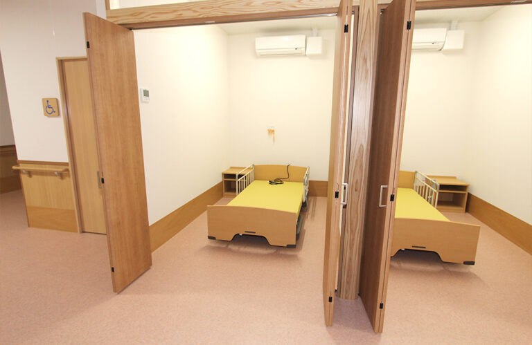 木製ベッドと収納が配置された介護施設の居室。明るく清潔な空間。